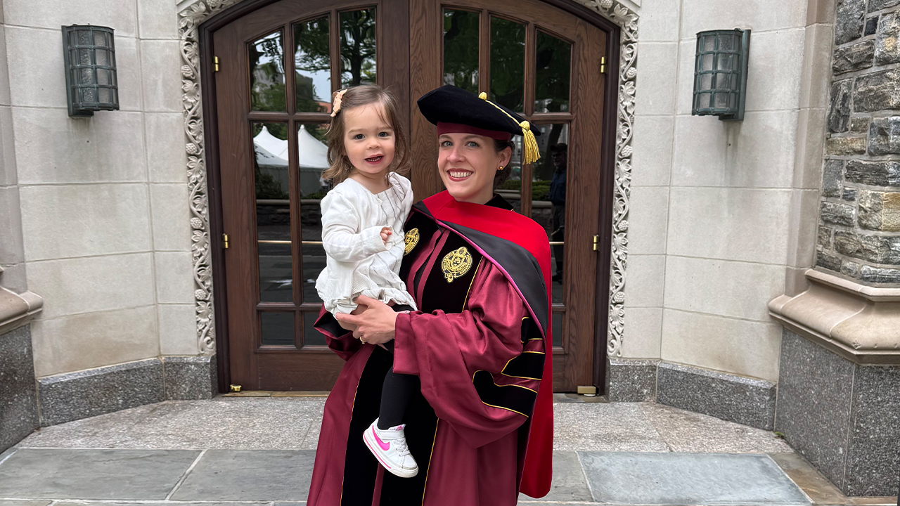 GSAS graduate Erica Restaino wearing cap and gown and holding daughter.