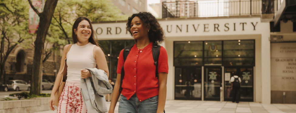 Two smiling students walk and laugh together on the plaza in front of the Fordham University Lincoln Center campus entrance.