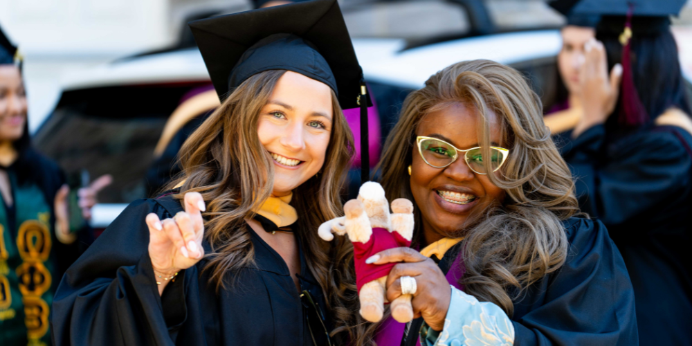 two students stand smiling in their caps and gowns. One student holds a stuffed ram, while the other makes a