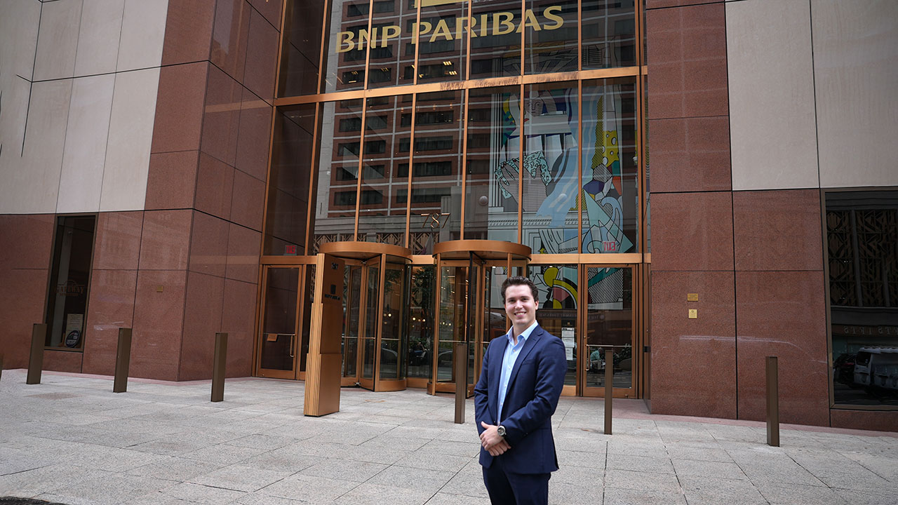 A student wearing a suit standing in front of an office building.