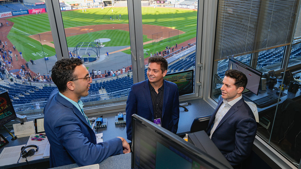 Three people talk in a press box at Yankee Stadium