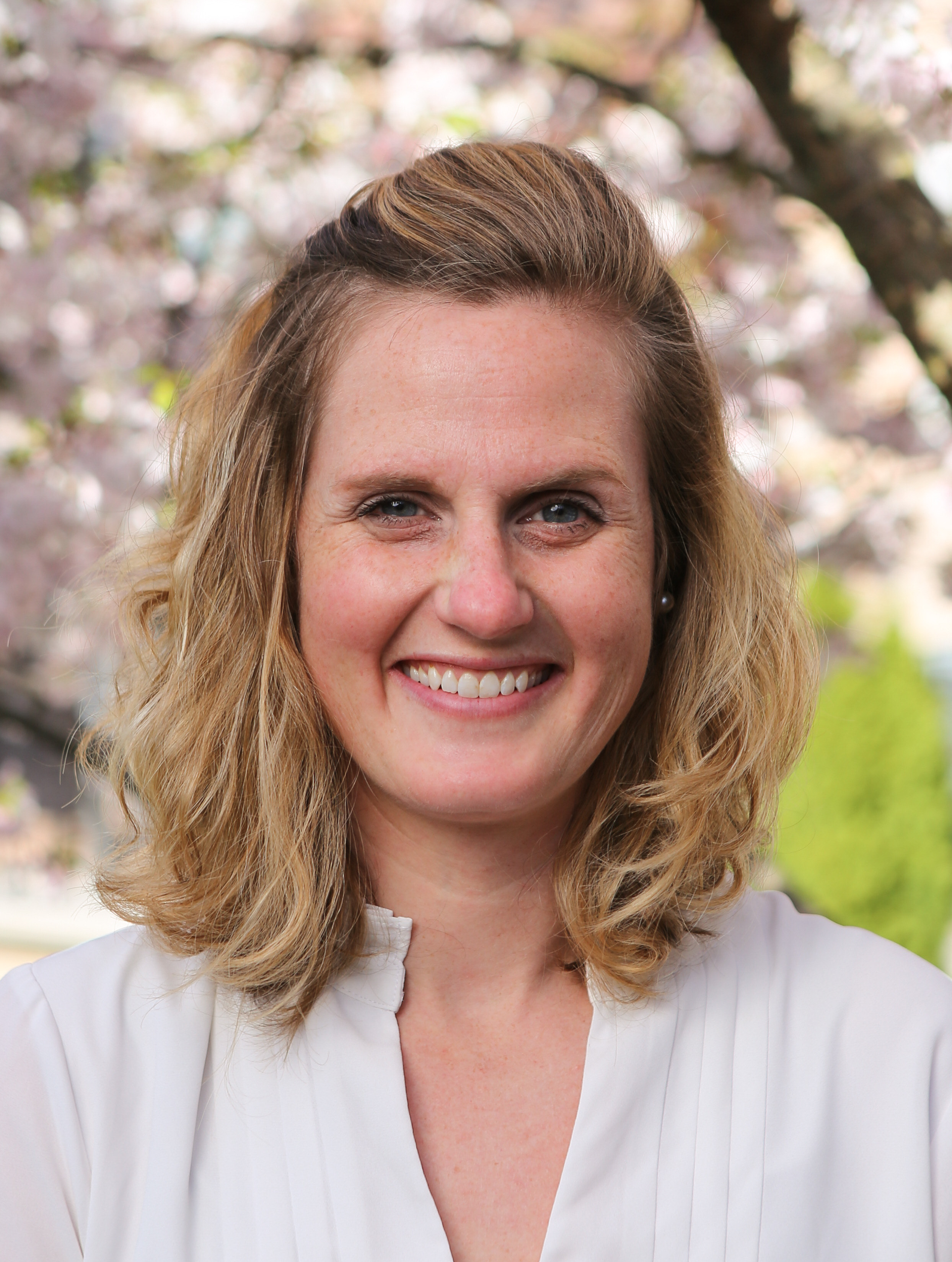 A headshot of Mara Brecht, a 2011 PhD graduate of Fordham's Theology Department. Brecht is wearing a white blouse in front of a background of tree blossoms.